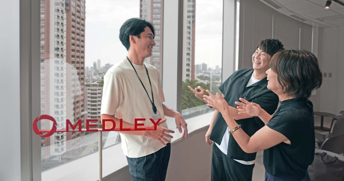 六本木ヒルズのメドレーオフィスの広い窓の前で、笑いながら話す三人。津野瀬さんとGaji-Labo横田のふたりが笑顔で小山さんを指さしている