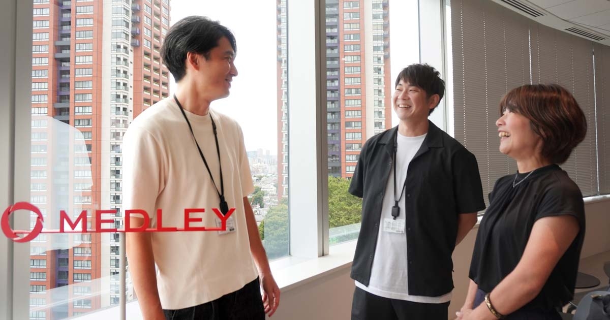 六本木ヒルズのメドレーオフィスの広い窓の前で、小山さんと津野瀬さんと笑顔で話すGaji-Labo横田